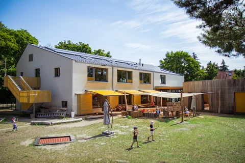 Modernes Kindergartengebäude mit Solardach, gelben Markisen und spielenden Kindern auf einem begrünten Außenbereich.