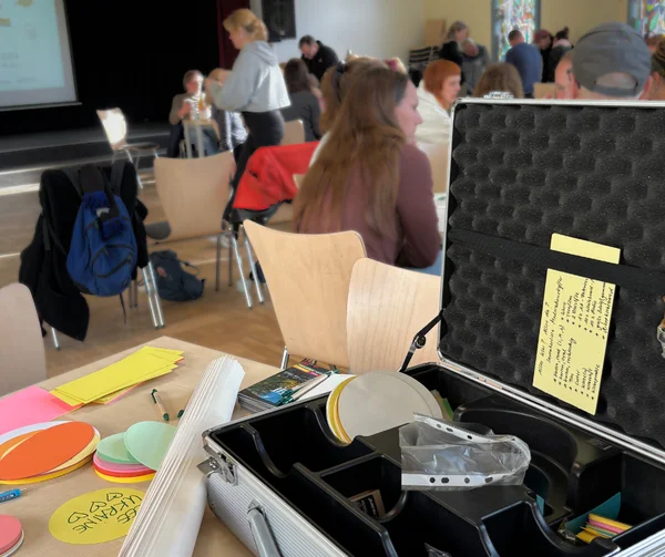 Ein Tisch voller Bastelmaterialien und ein geöffneter Koffer in einem Raum, in dem Menschen an Tischen sitzen. Ein Herz zeigt 'LOVE UKRAINE'.