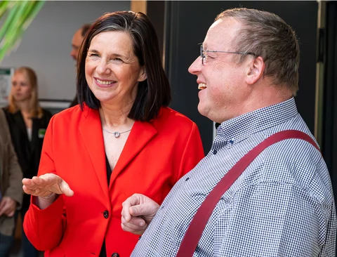 Eine Frau im roten Hosenanzug mit dunklen Haaren steht neben einem Mann mit kariertem Hemd und Hosenträgern. Beide lachen freundlich.