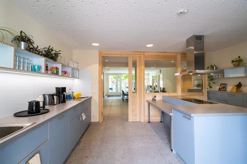 Eine helle, moderne Küche mit hellblauen Schränken, grauen Arbeitsplatten und strukturierter weißer Decke, mit Holzdurchgang zu einem Raum.
