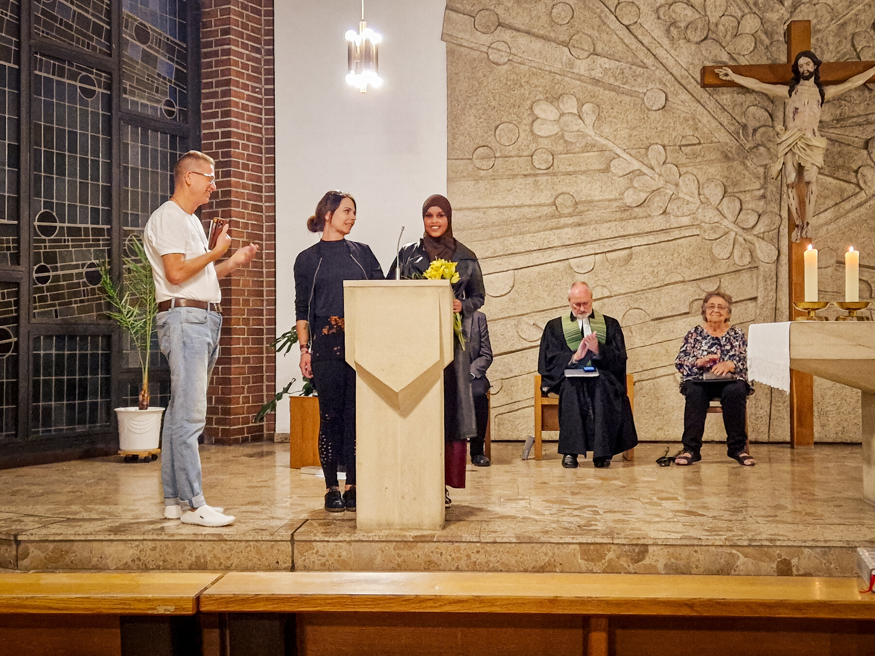 Diverse Gruppe bei einer Präsentation in einer Kirche. Eine Frau mit Hidschab hält Blumen, während ein Mann spricht.