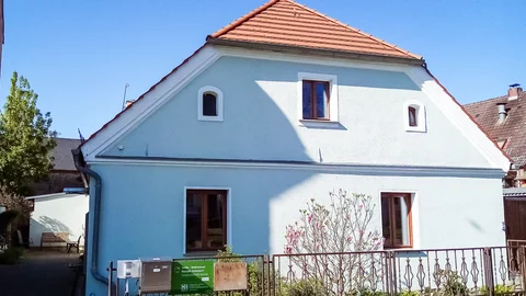 Hellblaues Haus mit rotem Ziegeldach, braunen Fenstern und Zaun mit Pflanzen. Ein Schild für sozialpsychiatrische Assistenz ist sichtbar.
