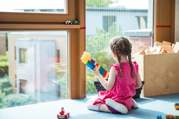 Ein Mädchen mit Zöpfen in einem rosa Kleid sitzt auf blauem Boden am Fenster, hält bunte Bausteine und schaut hinaus.