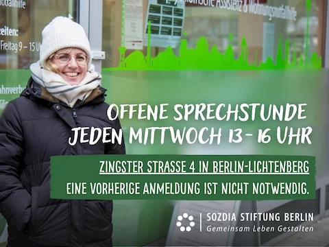 Frau mit Brille, weißer Mütze und Schal steht lächelnd vor einer Glaswand mit grünem Stadt-Silhouetten-Aufkleber und Text zu offener Sprechstunde in Berlin-Lichtenberg