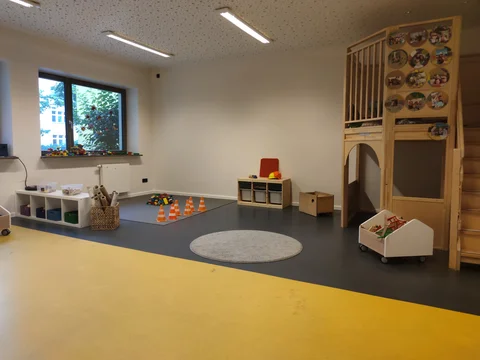 Ein heller Kinderspielbereich mit großem Fenster, Regalen, Spielzeug, einem Holzspielhaus mit Fotos und gelb-grauem Boden.