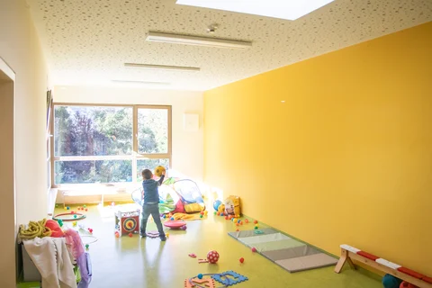 Ein Kind hält einen gelben Ball in einem farbenfrohen, sonnigen Spielzimmer mit Spielzeug auf grünem Boden und gelber Wand.