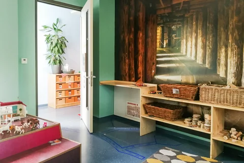 Ein farbenfroher Spielraum mit Wandgemälde eines Holztunnels, Spielzeugregalen, Tierfiguren und dunkelblauem Boden.