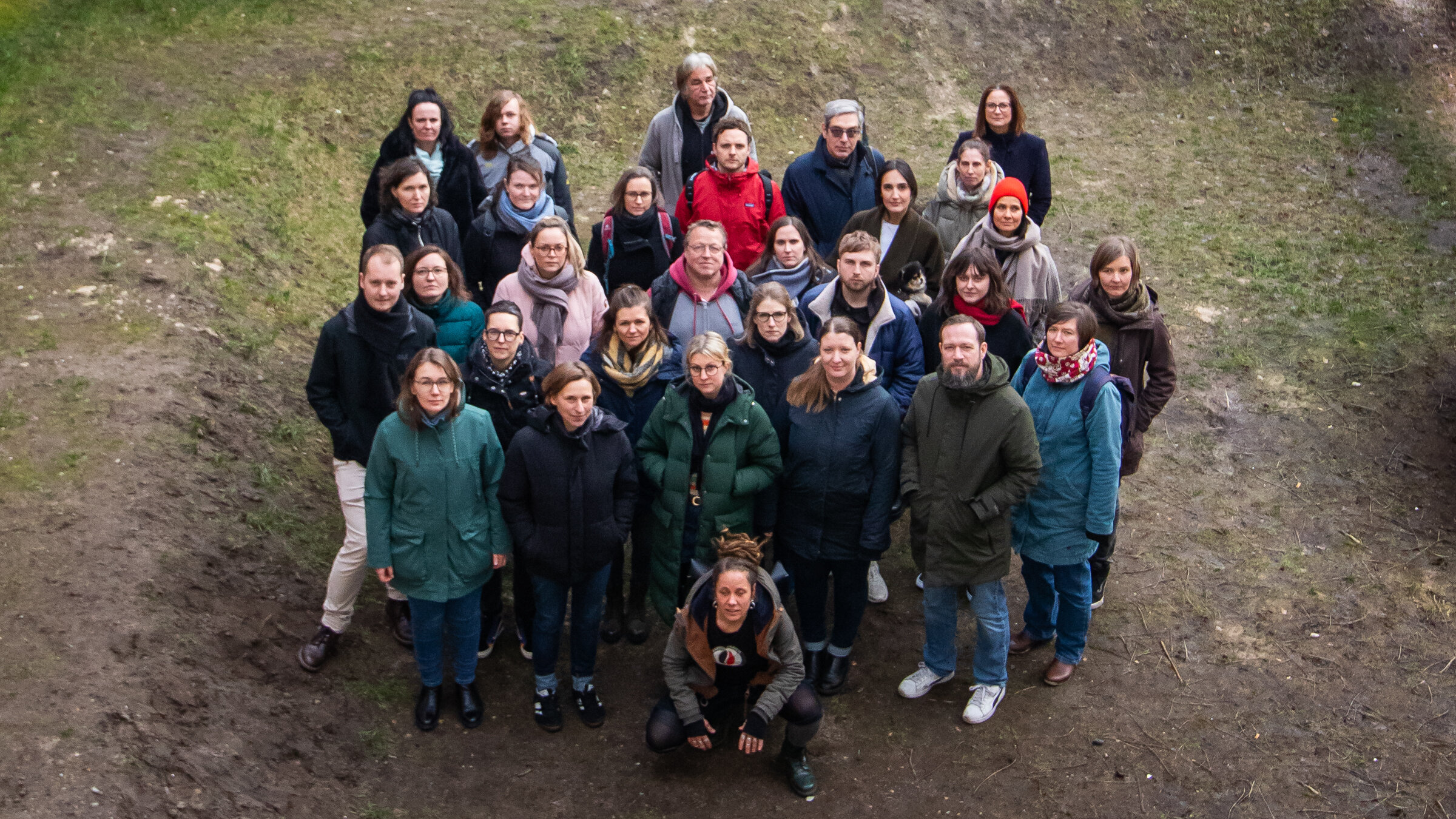 Eine große Gruppe vielfältiger Menschen steht eng zusammen auf einem grasbewachsenen, schlammigen Hang für ein Gruppenfoto.
