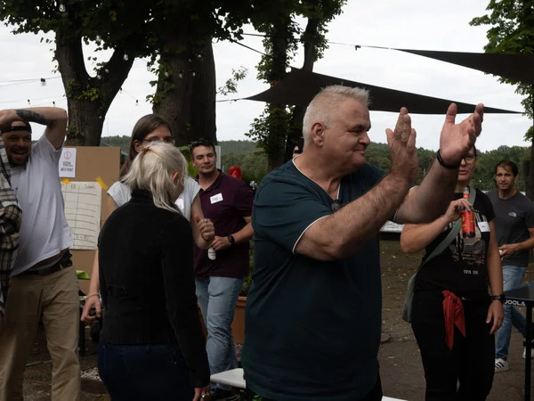 Mehrere Menschen stehen im Freien unter Bäumen, ein Mann im Vordergrund klatscht, andere schauen zu oder unterhalten sich.