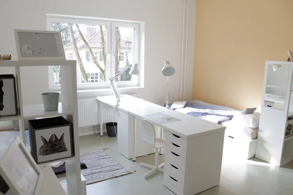 Ein helles, minimalistisches Zimmer mit weißem Schreibtisch am Fenster, einem Bett mit hellblauer Bettwäsche und weißen Regalen. Wände sind weiß und hellorange.