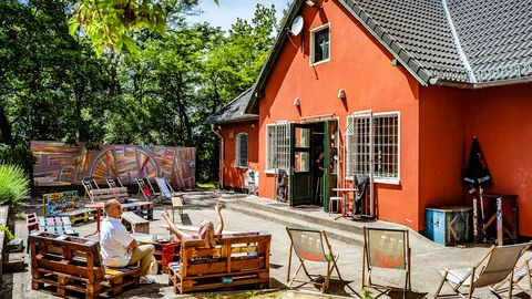 Outdoor-Café mit Menschen auf Palettensitzgelegenheiten, einem orangefarbenen Gebäude und bunter Graffitiwand unter Bäumen.