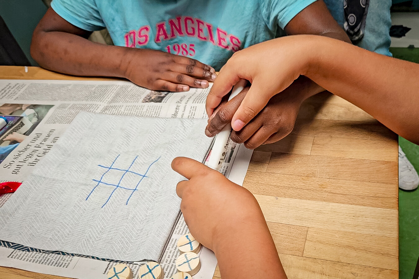 Hände von zwei Kindern spielen Tic-Tac-Toe auf Küchenrolle mit blauem Raster und hölzernen X-Steinen.