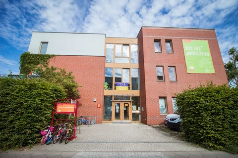 Vorderansicht der Kindertagesstätte Kita Vier Jahreszeiten mit roter Ziegelfassade, Glasfenstern, grünem Banner, Fahrradständer und Büschen.