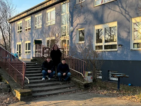 Vor einem zweistöckigen, blau gestrichenen Gebäude mit mehreren weißen Fenstern stehen drei Personen auf einer breiten Treppe. Zwei Personen sitzen auf den unteren Stufen, eine steht dahinter. Die Treppe ist von einem rotbraunen Geländer eingefasst. Im Hintergrund sind kahle Bäume und der Schattenwurf der Äste auf der Fassade zu sehen. Die Szene ist von Tageslicht erhellt.