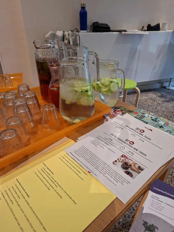 Mehrere Glaskrüge mit Wasser, Gurken und Früchten stehen neben bereitgestellten Trinkgläsern und Rezeptblättern.