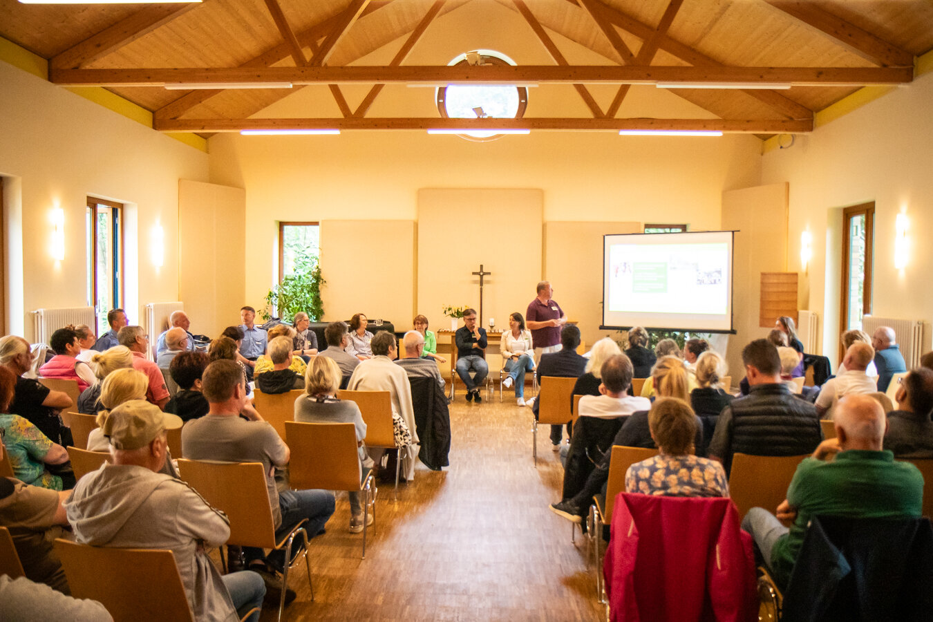 Zuhörer in Reihen lauschen einem Podium und Präsentator mit Projektionswand bei einer Veranstaltung in einem Raum mit Holzdach.