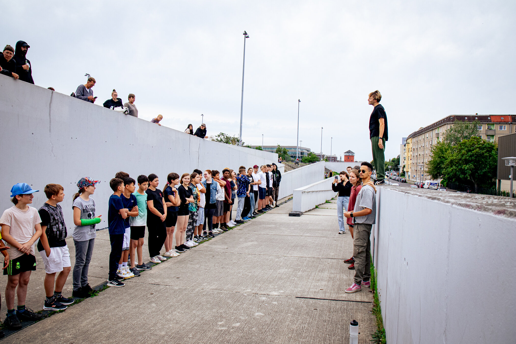 Zwei Reihen unterschiedlicher junger Leute stehen sich im Freien auf einem Betonweg gegenüber, ein Mann auf einer Mauer beobachtet sie.