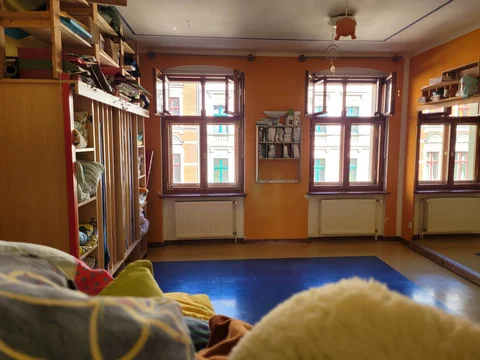 Helles, gemütliches Spielzimmer mit orangefarbenen Wänden, großen Fenstern, blauer Bodenmatte und Regalen.