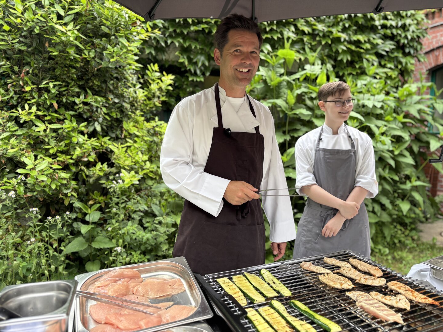 Zwei Personen in Kochkleidung stehen an einem Grill mit verschiedenen Fleisch- und Gemüsestücken, im Hintergrund grüne Pflanzen und eine Backsteinmauer