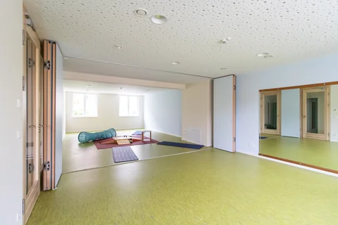 Heller Kinderspiel- oder Therapieraum mit hellgrünem Boden, Fenstern und Spiegel, ausgestattet mit Spiel- und Bewegungselementen.