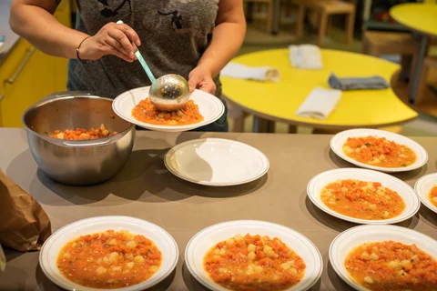 Mehrere Teller mit Gemüsesuppe auf einem Tisch, eine Person schöpft Suppe mit einer Kelle auf einen Teller.