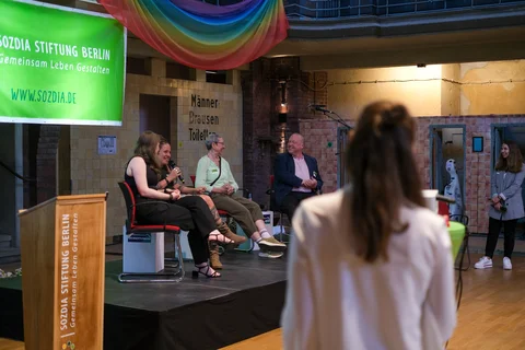 Auf einer Bühne sitzen vier personen. Im Vordergrund steht eine Frau mit langen Haaren.