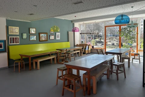 Heller Gemeinschaftsraum mit Holztischen, grüner Sitzbank, Wandkunst und großen Fenstern zu einem Spielplatz.