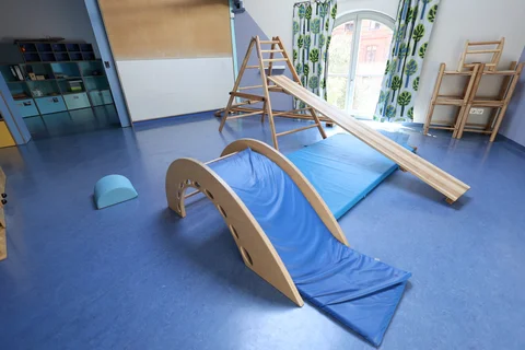 Ein helles Spielzimmer mit blauem Boden, ausgestattet mit hölzernen Klettergeräten, einer Rutsche, einem Wippbogen und blauen Weichbodenmatten.