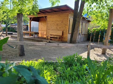 Sonniger Spielplatz mit Holzschuppen, Bank, Sandboden, grünem Bewuchs im Vordergrund und Bäumen. Ein Zaun quert die Mitte.