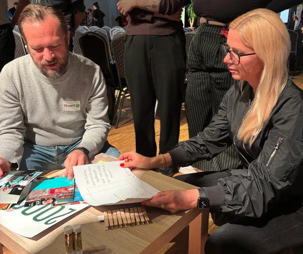 Eine Frau und ein Mann knien vor einem kleinen Tisch. Vor ihnen liegen Bilder und Klebstift.