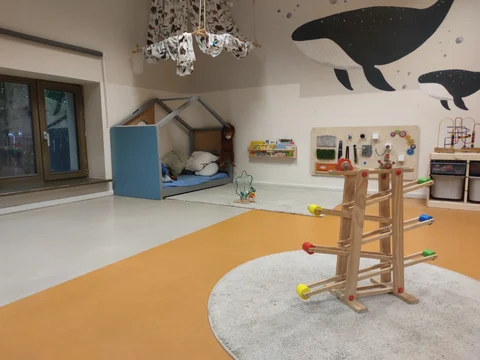 Helles Spielzimmer mit Hausbett, Holzspielzeug wie einer Kugelbahn und Motorikwand, ergänzt durch ein Wal-Wandgemälde.