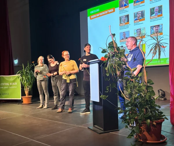 Fünf menschen stehen auf einer Bühne in der Hand eine Blume. Einer spricht ins Mikrofon.