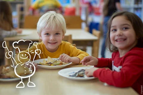 Zwei Kinder essen Bio Essen in einer SozDia Kita. Das Mädchen lacht in die Kamera und trägt einen roten Pullover. Der Junge ist blond und hat ein gelbes Shirt an. Sie sitzen vor ihrem Mittagessen.