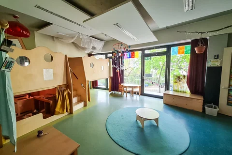 Ein heller, moderner Kinderspielraum mit großer Holzspielstruktur, rundem Teppich und großen Fenstern mit Blick auf Bäume.