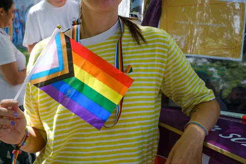 Person in gelb-weiß gestreiftem T-Shirt hält eine Regenbogen-Pride-Flagge mit zusätzlichen schwarzen, braunen und hellblauen Streifen
