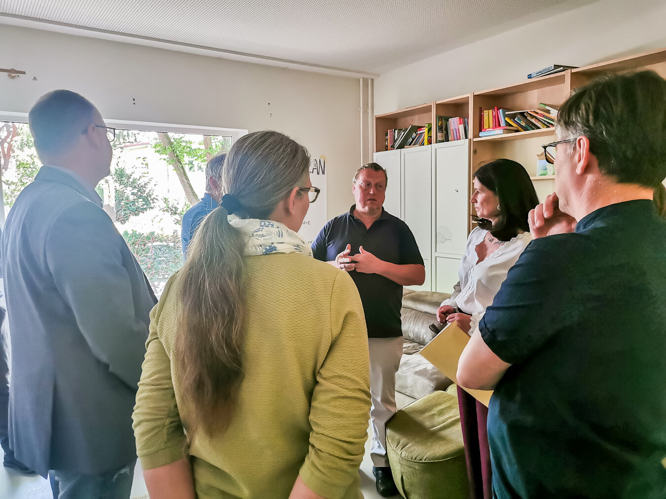 Eine Gruppe von Personen in einem informellen Meeting, versammelt in einem Raum, mit einem sprechenden Mann und aufmerksamen Zuhörern.