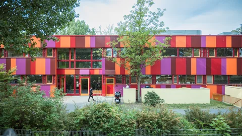 Das Bild zeigt die bunte Fassade eines zweistöckigen Gebäudes. Die Fassade ist mit vertikalen Streifen in lebhaften Farben wie Rot, Orange, Lila und Pink gestaltet. Die großen Fensterfronten sind symmetrisch angeordnet und lassen viel Tageslicht ins Innere des Gebäudes. Im unteren Bereich befinden sich mehrere Eingänge mit roten Rahmen. Links und rechts vom Gebäude sind Büsche und Bäume, die den Außenbereich umrahmen und für Grün sorgen. Ein Baum in der Mitte des Bildes steht vor dem Gebäude und spendet Schatten. Auf dem Gehweg vor dem Gebäude sind eine Person und ein Roller zu sehen. Der Außenbereich wirkt freundlich, lebendig und einladend, ideal für Kinder.