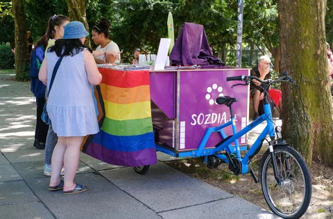 Dreirädriges Lastenfahrrad mit lila Kiste und Regenbogenfahne, davor stehen mehrere Personen auf einem Gehweg unter Bäumen
