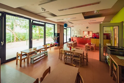 Heller Kindergarten-Gruppenraum mit Kindermöbeln, Küche und großen Fenstern zu einem grünen Außenbereich.