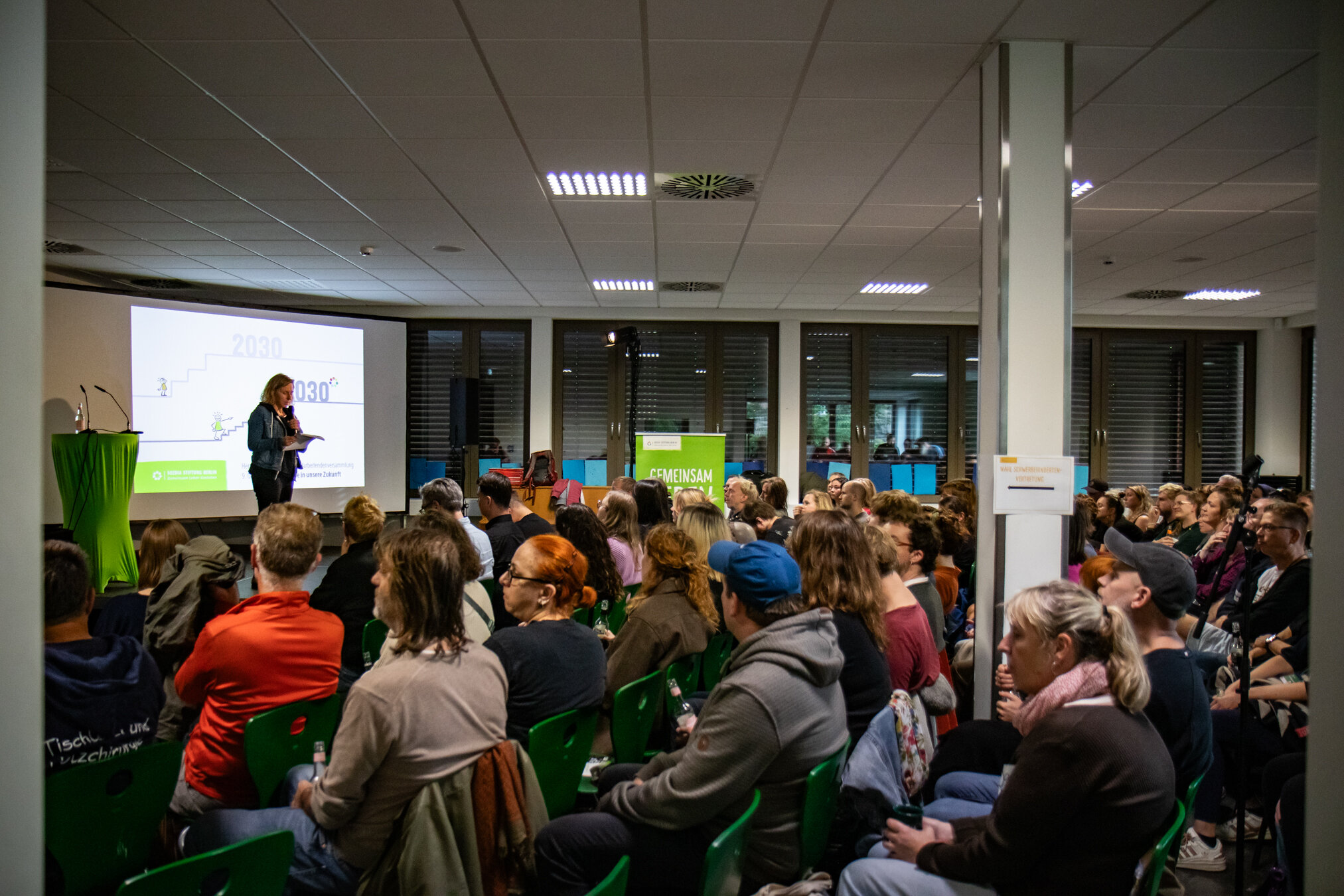 Eine Sprecherin präsentiert vor einem vielfältigen Publikum in einem Konferenzraum mit Leinwand "2030" und "GEMEINSAM"-Banner.