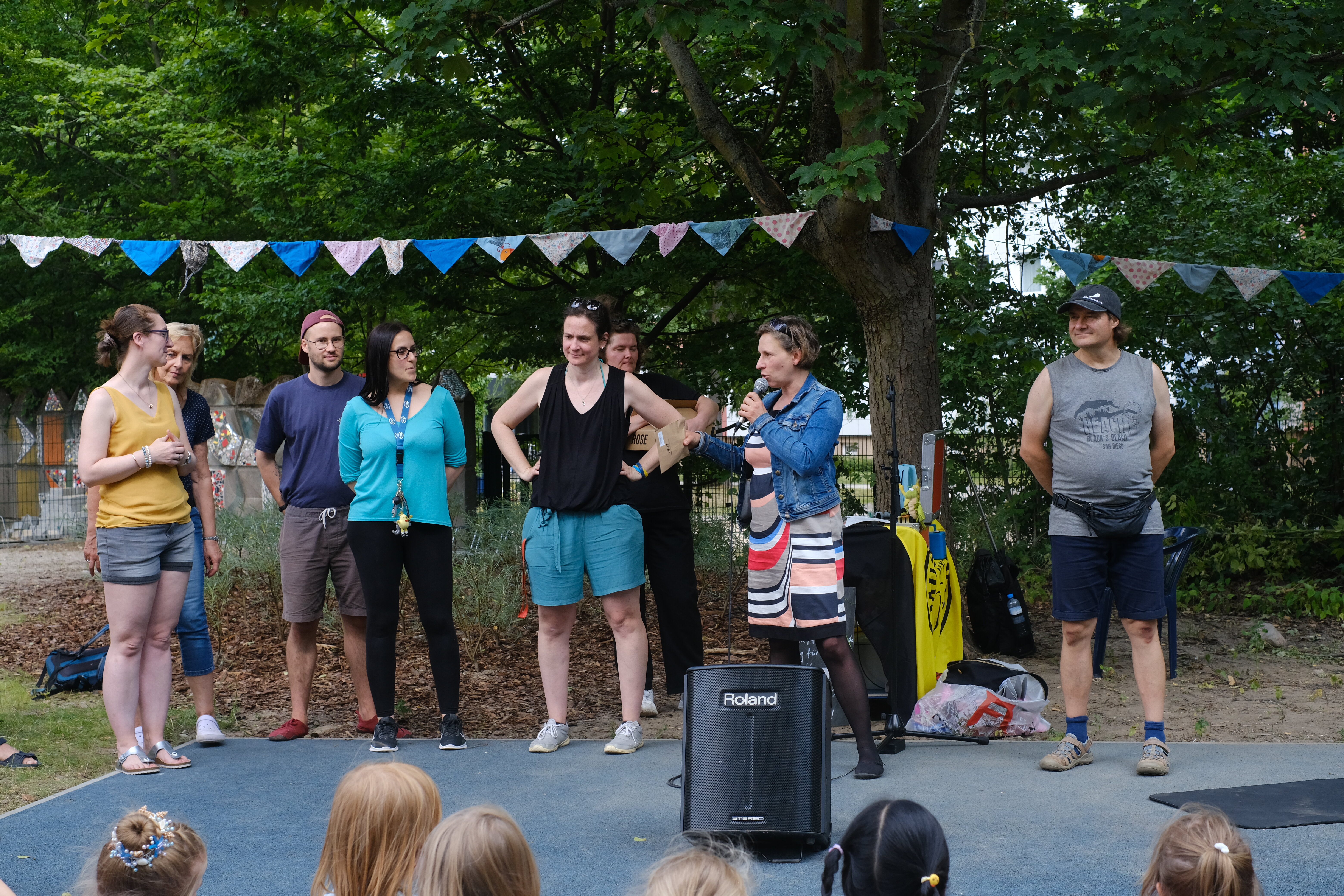 Eine Frau spricht in ein Mikrofon auf einer Outdoor-Bühne vor einer Gruppe Erwachsener und Kindern mit Bäumen im Hintergrund.