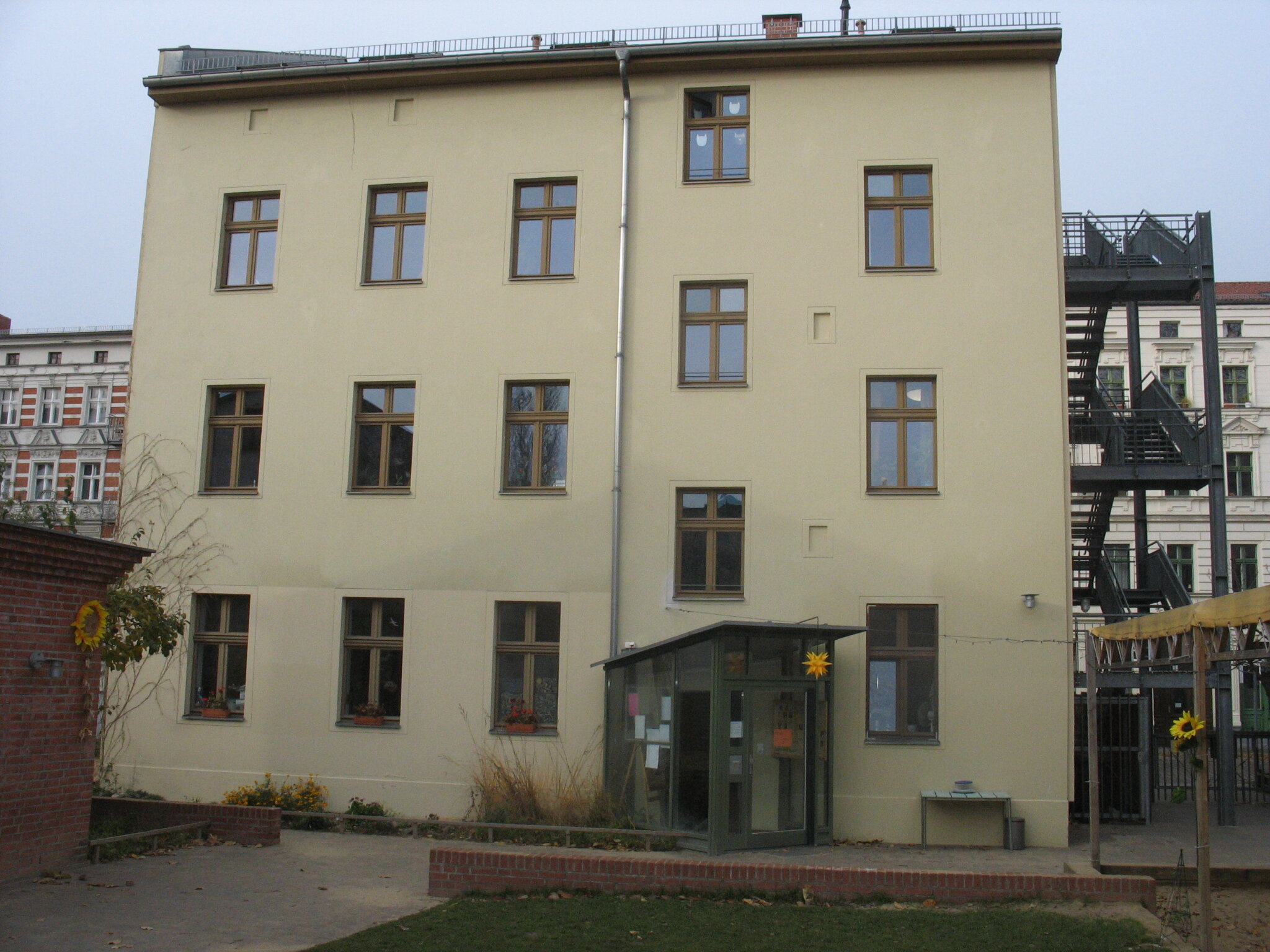 Hellgelbes, mehrstöckiges Gebäude mit braunen Fenstern, gläsernem Eingang und einer Metall-Feuerleiter an der Seite.