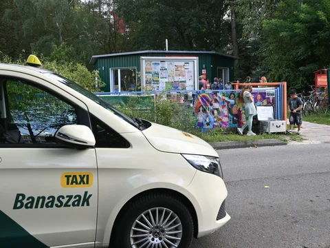 Taxi mit der Aufschrift 'Banaszak' steht an einer Straße vor einem umzäunten Bereich mit bunten Plakaten und mehreren Personen im Hintergrund
