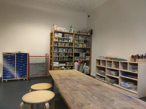 Heller Kunstraum mit großem, farbverschmiertem Holztisch, zwei Hockern und Regalen voller Bastelmaterialien.
