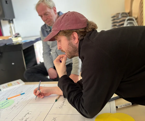 Ein Mann mit Kappe schreibt auf großem Rasterpapier während eines Workshops, ein weiterer Mann beobachtet im Hintergrund.