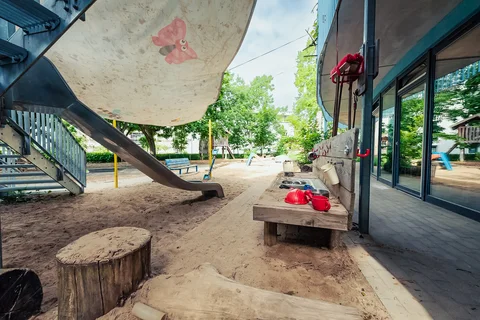 Ein Kinderspielplatz mit Rutsche, großem Sandkasten, einer Outdoor-Spielküche und einem Sonnensegel mit Schmetterling an einem sonnigen Tag.