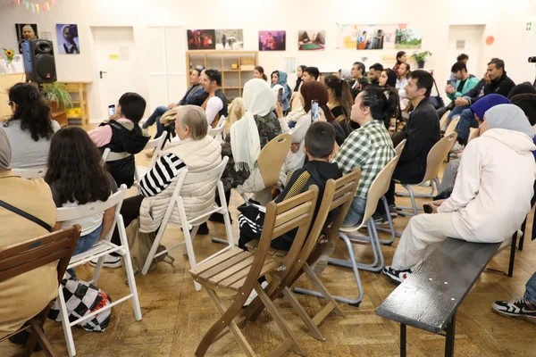 Eine vielfältige Gruppe von Menschen, darunter Erwachsene und Kinder, sitzt in einem Gemeindesaal und schaut aufmerksam nach vorne.