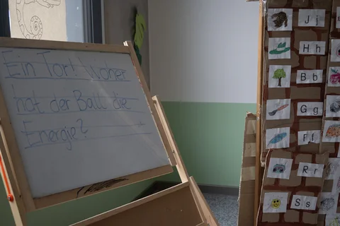 Kindertafel mit der handschriftlichen Frage 'Ein Tor! Woher hat der Ball die Energie' und daneben eine Wand mit Buchstaben- und Bildkarten.