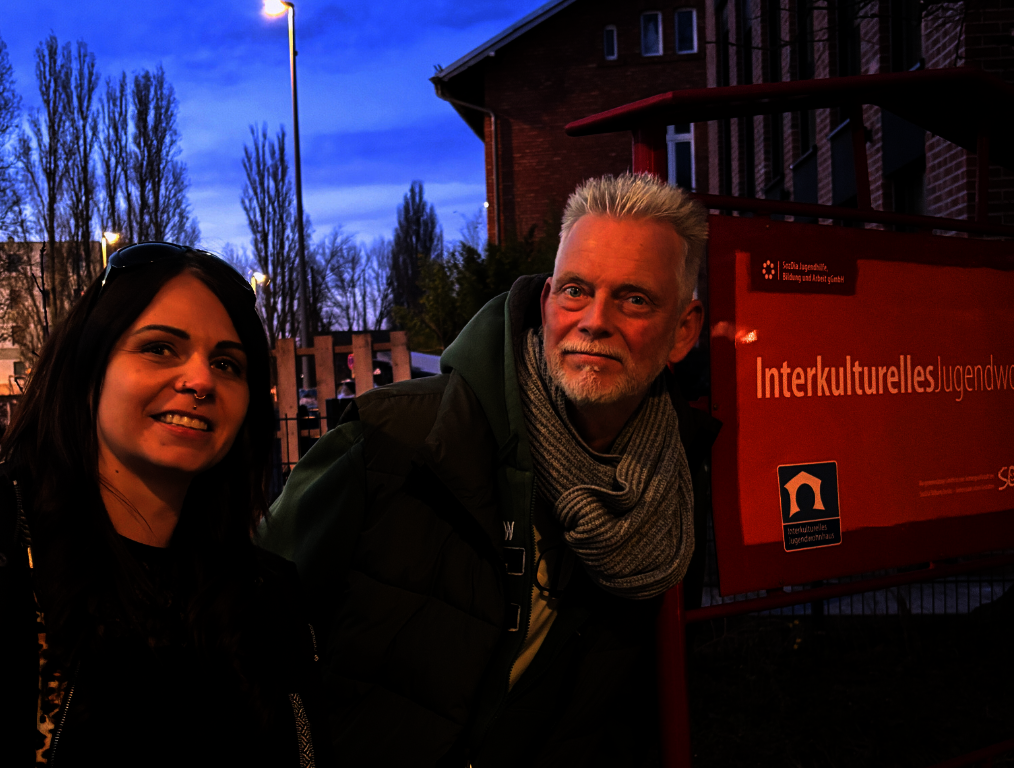Oskar hat graue Haare und Maike lange dunkle Haare. Sie stehen in der Dämmerung vor einem orangenen Schild auf dem steht "Interkulturelles Jugendhaus".