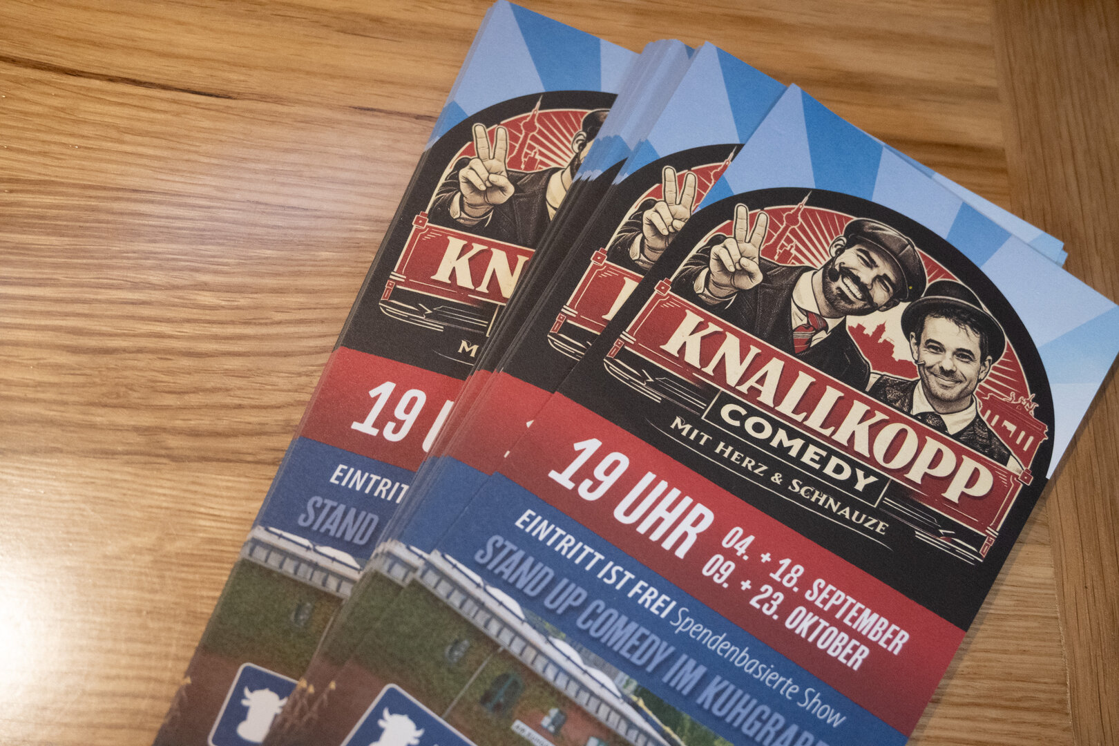 Zwei Flyer für die Stand-up-Comedy-Veranstaltung "Knallkopp Comedy" mit zwei Männern, die jeweils ein Peace-Zeichen zeigen, auf einem Holztisch